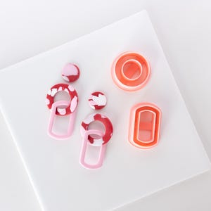 Puede incluir: Un par de pendientes de declaración rosa y rojo con un diseño geométrico. Los pendientes están hechos de arcilla polimérica y tienen un efecto marmoleado. Los pendientes se muestran sobre una superficie blanca con otras formas de arcilla polimérica.