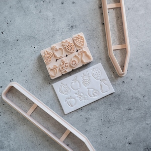 May include: A set of light peach-coloured tools including two elongated pencil-shaped cutters, a rectangular silicone mould with eight fruit designs, and eight individual small square stamps featuring matching fruit motifs like strawberries and pears.