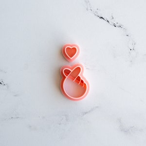 May include: Two pink plastic cookie cutters, one shaped like a heart and the other shaped like a hand making a heart sign.
