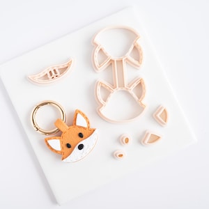 May include: A collection of fox-themed craft supplies, including a finished felt fox keychain with a gold ring, and several light pink plastic cookie cutters. The cutters are in the shape of a fox head, ears, and other fox features.