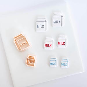 Pode incluir: Dois cortadores de biscoitos de plástico cor de pêssego em forma de caixa de leite e seis formas brancas de caixa de leite com a palavra MILK impressa em tinta cinza, vermelha e azul. As formas estão dispostas em pares, mostrando os cortadores.