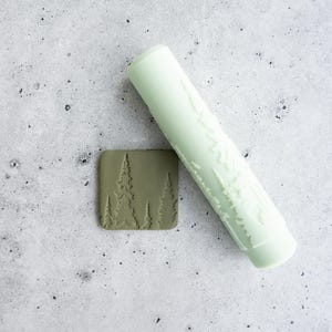 Può includere: Un mattarello cilindrico color verde menta pallido con un motivo a foresta in rilievo accanto a una piastrella quadrata in argilla verde oliva che presenta lo stesso disegno di alberi, mostrando l'impronta lasciata dal rullo.