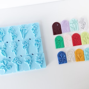 May include: A set of twelve light blue rectangular silicone molds featuring various floral line art designs, alongside twelve finished arch-shaped pieces in assorted solid colors, each displaying a matching embossed floral pattern.