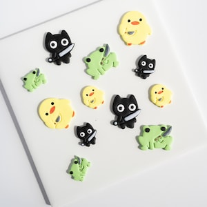 May include: A collection of small, flat, decorative figures made of polymer clay. The set includes black cats, light green frogs, and yellow chicks, each holding a small silver-colored knife. The figures are arranged in a scattered pattern.