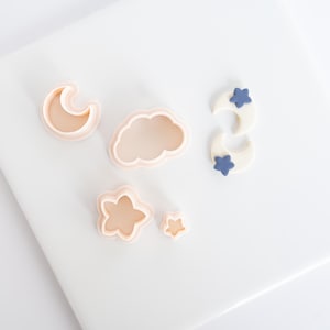 May include: A collection of light peach-coloured plastic cookie cutters in the shapes of a crescent moon, a cloud, and two flowers of different sizes, alongside two white crescent moon shapes decorated with small blue star accents.