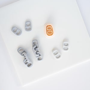 May include: A collection of small, oval-shaped soda can tab replicas. There are several light gray individual tabs, two short chains made of linked gray tabs, and one single tab featuring a light peach border with a wood-grain textured center.