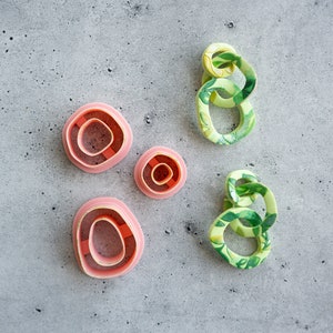 May include: Four pink and green plastic cookie cutters in different shapes and sizes. The cutters are on a gray textured surface.