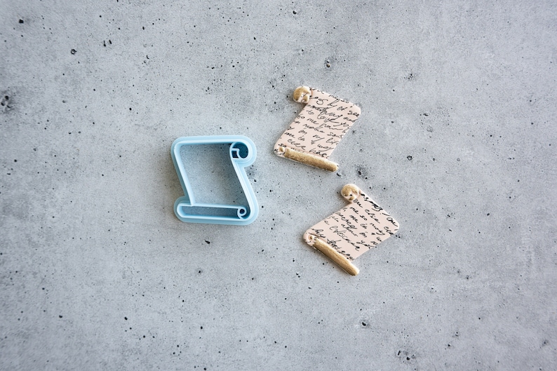 May include: A blue plastic cookie cutter shaped like a scroll, next to two fondant scrolls with gold accents and a handwritten script design.