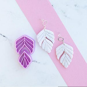 May include: A pair of white speckled leaf-shaped earrings with silver-toned circular connectors and a matching purple plastic cutter tool featuring a segmented leaf design. The earrings consist of two stacked leaf segments with a textured finish.