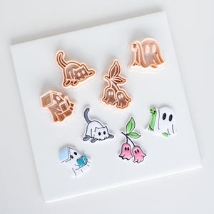 Puede incluir: Un conjunto de cortadores de galletas de plástico y sus correspondientes recortes de fondant o arcilla de colores. El set incluye formas de gato, fantasma, casa y flores en forma de campana, con contornos detallados y colores vibrantes.