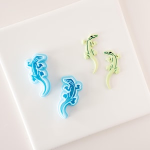 May include: Four plastic cookie cutters in the shape of lizards. Two are light blue and two are light green.