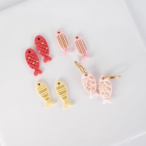 May include: A collection of fish-shaped earrings and charms in various colors. The earrings and charms are in red, pink, and yellow with gold accents. Some have scale or stripe patterns, while others are on gold hoops.