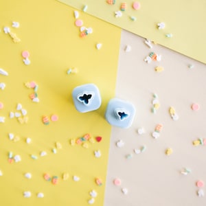 Puede incluir: Dos cortadores de galletas de plástico azul en forma de mariposa sobre un fondo amarillo y beige con chispas de colores pastel.