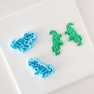 May include: Three alligator cookie cutters, two are light blue and one is green. The cutters are on a white surface.