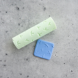 Pode incluir: Um rolo de massa cilíndrico verde-claro e um pequeno azulejo quadrado azul, ambos com um padrão em relevo de pássaros a voar. O rolo é alongado, enquanto o azulejo é plano e compacto, exibindo um motivo aviário consistente.