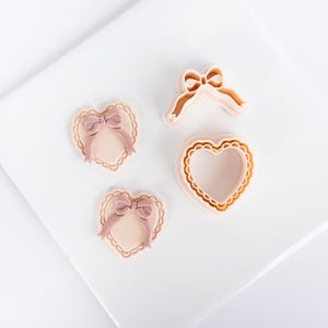 Può includere: Quattro formine per biscotti e decorazioni a forma di cuore. Due decorazioni a forma di cuore con fiocchi rosa e bordi smerlati. Due formine per biscotti a forma di cuore, una con un fiocco e una con un bordo smerlato. Tutti gli articoli sono in tonalità rosa e pesca.