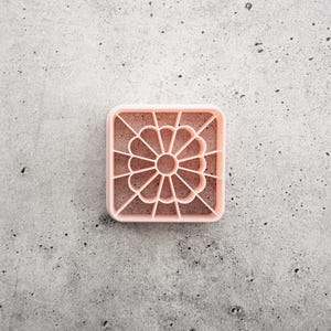 May include: A square-shaped light pink cookie cutter featuring an intricate floral design inside. The cutter has a central circular element surrounded by twelve radiating petals, all enclosed within a square frame with straight lines extending to the corners.