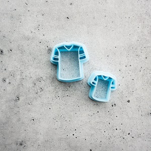 May include: Two blue plastic cookie cutters shaped like football jerseys. The larger cutter is in the background and the smaller cutter is in the foreground.