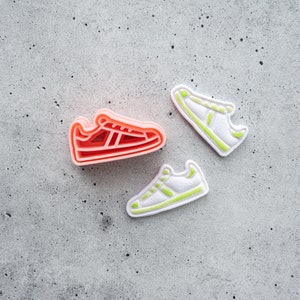 May include: Three cookie cutters shaped like trainers. One is pink, one is white with green trim, and the other is white with yellow trim.