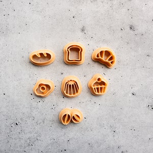May include: Set of seven plastic cookie cutters in various shapes, including a loaf of bread, a slice of bread, a croissant, a cupcake, a jelly mold, an egg, and a pair of coffee beans. The cutters are a light brown color and are arranged on a gray textured surface.