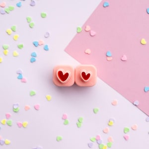 May include: Two square, light pink plastic stamps, each featuring a raised heart-shaped design in the center. The stamps are surrounded by a scattering of small, colorful heart-shaped confetti in shades of blue, yellow, green, and pink.
