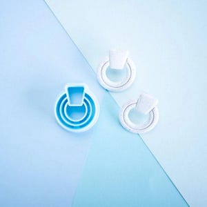 May include: A bright blue plastic ring-shaped cookie cutter alongside two white speckled clay earrings, each featuring a circular hoop design with a small trapezoidal top piece, arranged on a light blue surface.