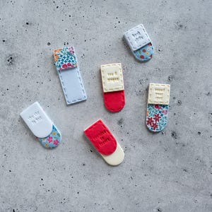 May include: Five magnetic bookmarks in various colors and patterns. One is white with floral fabric, one is red with a cream top, one is cream with floral fabric, one is white with blue text, and one is red with cream text. Each has stitched edges.