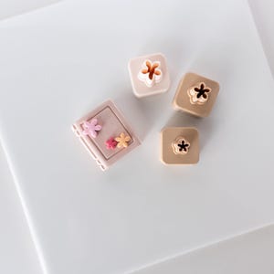 May include: A set of three square-shaped beige and tan clay cutters featuring flower-shaped cutouts, accompanied by a rectangular beige clay block decorated with three small, colorful flower-shaped embellishments in pink, red, and yellow.
