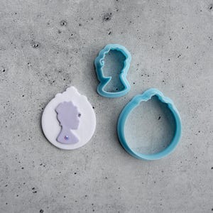 Könnte beinhalten: Drei hellblaue Ausstecher und ein weiß-lilafarbenes Cameo. Das Cameo hat eine lilafarbene Silhouette des Profils einer Frau mit einem Juwel. Die Ausstecher haben die Form eines Frauenprofils und eines dekorativen Ovals.