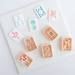 Puede incluir: Un conjunto de fichas de Mahjong en blanco, azul claro, verde, rosa y melocot&oacute;n. Las fichas presentan s&iacute;mbolos y dise&ntilde;os detallados, dispuestos sobre una superficie blanca. Las piezas del juego son rectangulares y cuadradas.