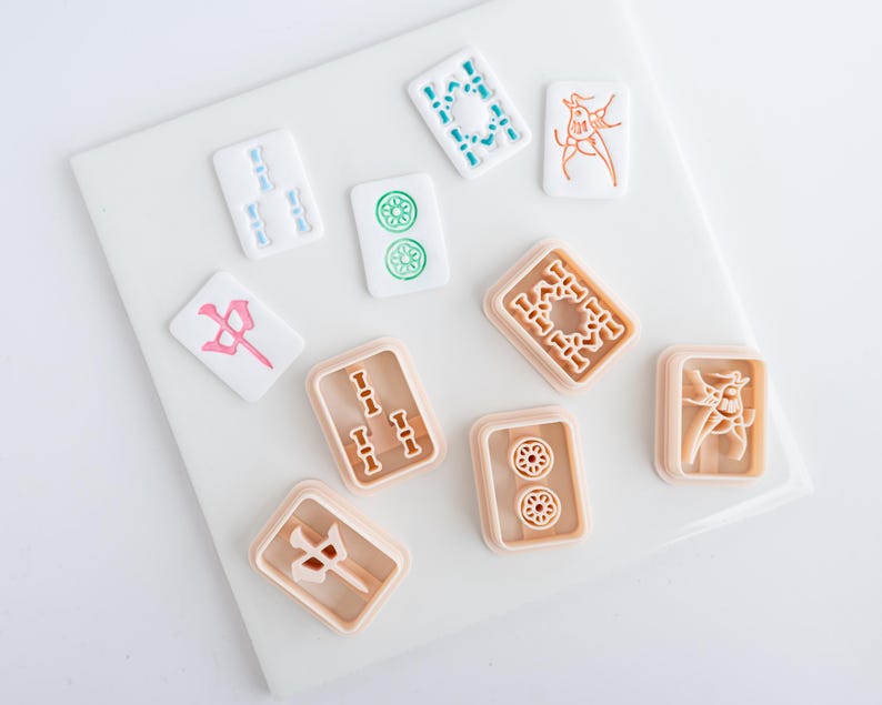 Puede incluir: Un conjunto de fichas de Mahjong en blanco, azul claro, verde, rosa y melocot&oacute;n. Las fichas presentan s&iacute;mbolos y dise&ntilde;os detallados, dispuestos sobre una superficie blanca. Las piezas del juego son rectangulares y cuadradas.