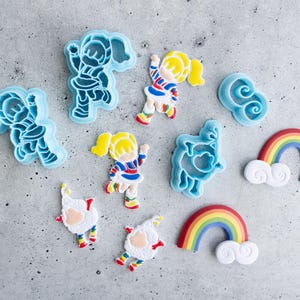 May include: A set of blue plastic cookie cutters featuring a rainbow, clouds, a sheep, and a girl with a rainbow dress.