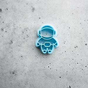 May include: A light blue plastic cookie cutter shaped like an astronaut. The design features a rounded helmet, a central rectangular chest panel, and simple limb outlines, creating a clear silhouette for cutting dough into a space-themed figure.