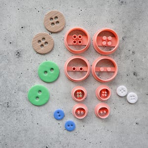 May include: A collection of various craft supplies including two tan ribbed circular buttons, two smooth green buttons, two small blue buttons, two tiny white buttons, and several peach-colored circular molds with internal four-hole patterns.