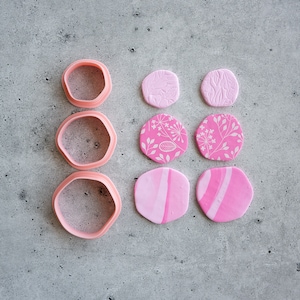 May include: Three pink plastic cookie cutters in different sizes and shapes, next to six pink clay circles with different patterns and textures.