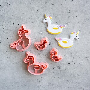 May include: A collection of pink plastic unicorn-shaped cookie cutters in two sizes alongside two finished unicorn-shaped cookies featuring white bodies, yellow inner rings, and colorful manes and tails, all arranged on a gray textured surface.