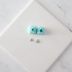May include: Two light blue square-shaped clay cutters featuring an ivy leaf design, accompanied by two small, light gray, star-shaped clay cutouts. The cutters have a raised central section that forms the leaf shape, while the gray pieces are flat.