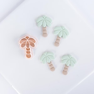 Pode incluir: Um cortador de biscoitos em forma de palmeira rosa claro ao lado de quatro pequenas figuras de palmeiras feitas de folhagem verde claro e troncos bege. O cortador apresenta um interior oco com um contorno em relevo do design.
