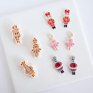May include: A collection of three light-colored plastic cookie cutters shaped like a nutcracker, a ballerina, and a mouse, alongside six matching colorful clay or resin charms featuring the same characters in red, pink, and gray tones.