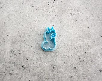 May include: A light blue plastic cookie cutter shaped like a llama with a small heart detail on its cheek. The cutter features a defined outline and an inner impression detail for the llama's facial features and ears, designed for cutting dough.
