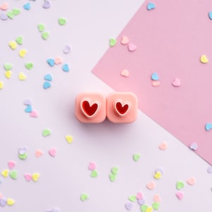 May include: Two pink heart-shaped cookie cutters with red hearts inside, on a white and pink background with pastel pink, blue, and yellow confetti hearts scattered around.