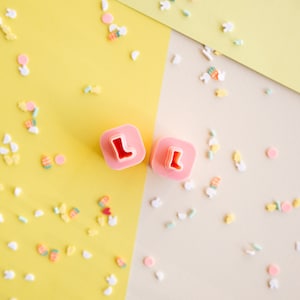 May include: Two pink plastic cookie cutters shaped like the letter L, on a background of yellow and beige with pastel colored sprinkles.