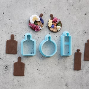 Può includere: Un set di sette stampini per biscotti, tra cui due forme di tagliere in legno marrone, uno stampino rotondo blu, uno stampino rettangolare blu e due taglieri marroni con vari condimenti.