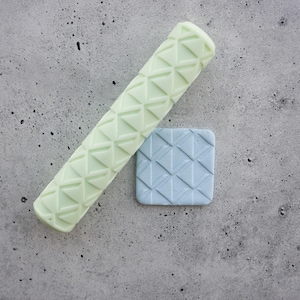 Pode incluir: Um rolo de massa cilíndrico verde-pálido com um padrão geométrico de triângulos em relevo ao lado de um pequeno azulejo quadrado azul-claro que exibe o mesmo design triangular gravado, ambos com uma textura repetitiva consistente.