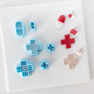 May include: A set of blue and red plastic cookie cutters shaped like a pill, a cross, and a bandage. The cutters are arranged on a white surface.