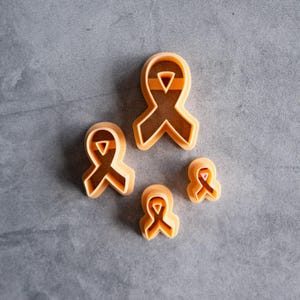 May include: A set of four light orange plastic cookie cutters, each shaped like an awareness ribbon. The cutters vary in size, with one large, one medium, and two smaller ribbons, all featuring a distinct cutout design in the center of the loop.
