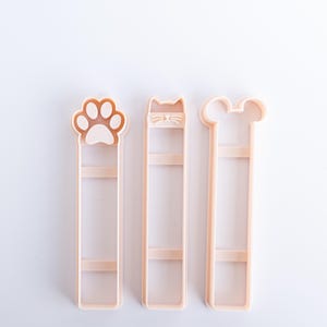 May include: Three light peach-colored plastic bookmark molds arranged vertically. The left mold features a paw print design, the center mold displays a cat face with whiskers, and the right mold has a rounded mouse ear shape at the top.