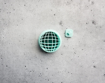 May include: A light mint green circular object featuring a grid pattern resembling a globe, accompanied by a small, separate square piece with a central protrusion. Both items are solid in color and display a smooth, matte finish throughout.
