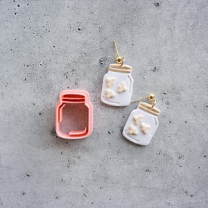 May include: A pink plastic jar-shaped cookie cutter alongside two white jar-shaped earrings with gold-tone hardware. Each earring features small, light-colored, textured shapes inside the jar design, suspended from gold-tone ball-post earring findings.