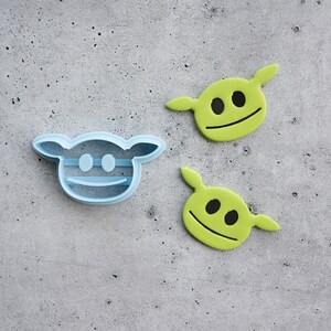 May include: A light blue plastic cookie cutter shaped like a cartoon head with large ears, alongside two lime green fondant or cookie cutouts featuring the same head shape with black circular eyes and a horizontal line for a mouth.